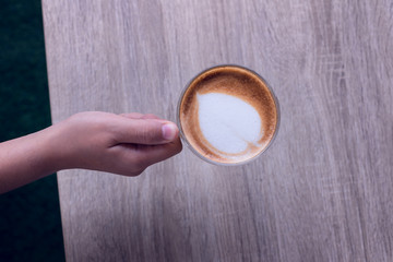 Female hands holding a cup of coffee with heart make up on face of milk cream on wooden table, top view