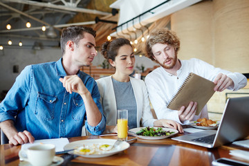 Clever student showing his sketch for coursework in notepad to groupmates during lunch break in cafe