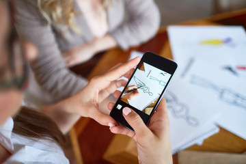 Close-up shot of unrecognizable woman photographing on smartphone while her talented friend making sketch, focus on foreground