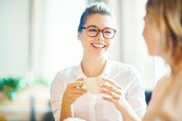 Head and shoulders portrait of attractive young woman with charming smile enjoying fragrant coffee while hanging out with best friend in cozy small cafe