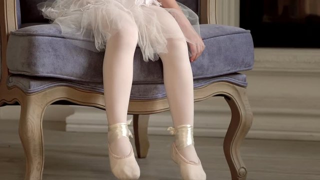 The little fair-haired girl sits on a chair at a fireplace in a white tutu. Legs of the little ballerina in white pointes. Legs close up