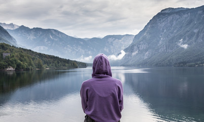 Solitary woman wearing purple hoodie watching tranquil overcast morning scene at lake Bohinj, Alps mountains, Slovenia.
