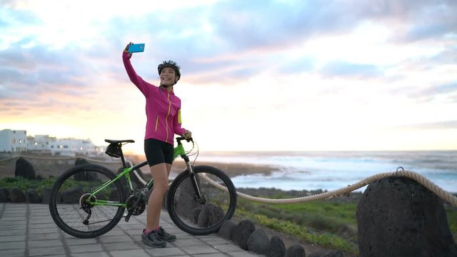 Woman cyclist biking taking selfie using smart phone while MTB mountain biking. Beautiful woman in sportswear is posing with bicycle. She is photographing using smart phone in nature with bike.
