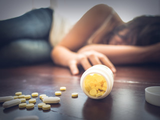 Woman taking medicine overdose and lying on the wooden floor with open pills bottle. Concept of overdose and suicide. - obrazy, fototapety, plakaty