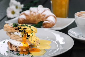 Poached eggs sandwich for breakfast with croissant, juice, coffee and chamomile on dark wood background