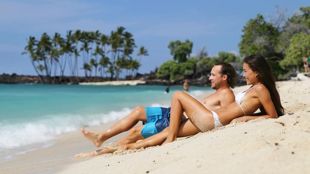 Vacation travel couple relaxing on beach talking laughing having fun. Happy multiethnic man and woman are lying on sand during summer vacation. They are enjoying on sunny day.