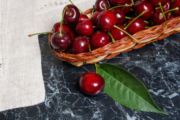 Several red sweet cherries and big green leaf on the table. Fresh organic cherry in yellow wooden basket on dark marble background..