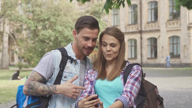 Attractive caucasian students taking selfies on campus. Cute young people using some smartphone app to make funny effects on images. Handsome brunette guy and his pretty female classmate grimacing for