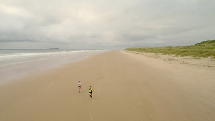 Fototapeta na wymiar Runners on Beach #1 - An aerial drone shot of a man and woman running on the beautiful beach of Bamburgh, North England at dawn. Dunes and waves are on either side. The runners run towards and under t