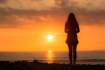 Young woman stands looking out to sea at the sunset