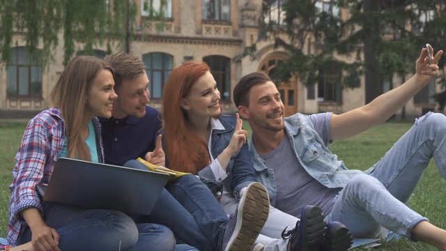Four cute caucasian students taking selfie on the lawn on campus. Attractive young people smiling for the smartphone camera. Pretty redhead girl making bunny ears behind brunette guy's head