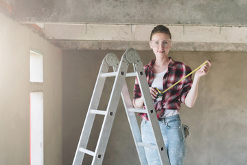 A builder, dressed in a plaid shirt and construction glasses, stands on a stepladder and holds...