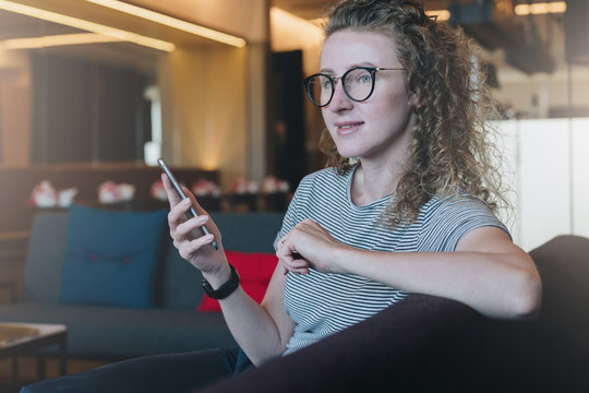 Young smiling curly business woman hipster in glasses sits on sofa in resting room, cafe and holds smartphone. Girl thinks