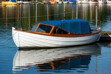 Small white wooden motorboat covered with blue tarp. Boat moored at wooden pier and have rope hanging over it.