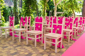 Fototapeta na wymiar Wedding chairs on each side of archway with pink cloth. Place for wedding ceremony decorated in pink color, wooden chairs for guests outdoors. Wedding ceremony in pink color