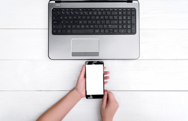 Top view on white wooden table with open blank laptop computer and girl's hands with cell phone, free space. Mobile phone with white screen, copy space