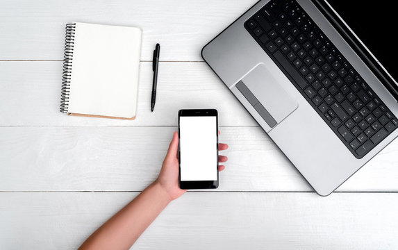 Top view on white wooden table with open laptop computer, cell phone in girl's hand and blank empty diary with pen, free space. Mobile phone with blank white screen, copy space