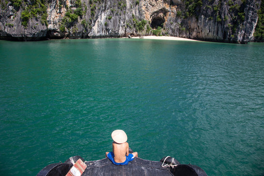 man wearing traditional vietnamese hat sitting in front of ship cruising among beautiful limestone rocks and secluded beaches in Ha Long bay, UNESCO world heritage site, Vietnam