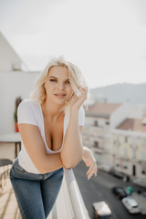 Young blonde woman sitting and relaxing at terrace on chair with city view. Fresh air in the morning of weekend or free day.