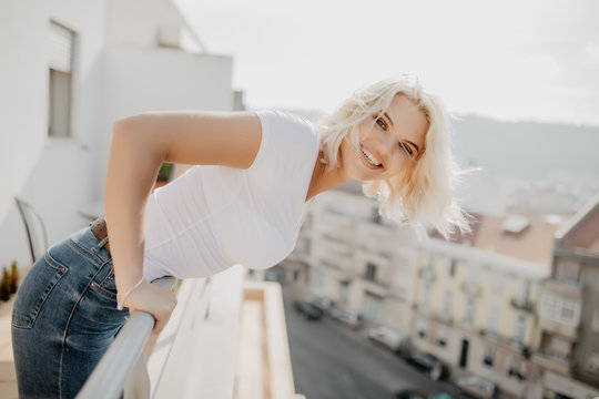 Young blonde woman sitting and relaxing at terrace on chair with city view. Fresh air in the morning of weekend or free day.