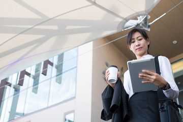 businesswoman holding coffee and digital tablet outside office building. beautiful young asian woman with tea go to work outdoors. female adult with disposable paper cup walking in business area.