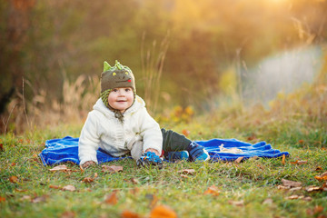 Little smiling boy sitting on a blue plaid and playing with toys on a green lawn at sunset