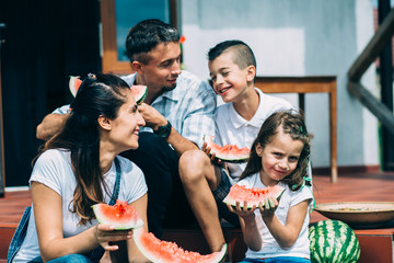 Close up of happy parents with two cute children sitting on the porch steps and eating watermelon in autumn time. New housing, parenthood and children concept.