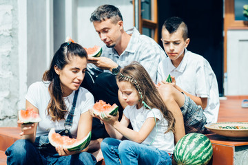 Close up of happy parents with two cute children sitting on the porch steps and eating watermelon in autumn time. New housing, parenthood and children concept.