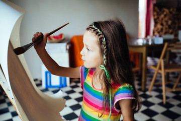 Cute little artist girl painting with a large brush picture in studio or home over floor with checkerboard texture. Education, creation, art, people and children concept.