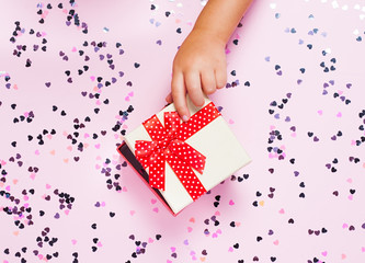 Child's hand opening gift box on pinh background with heart shaped confetti. Top view, flat lay