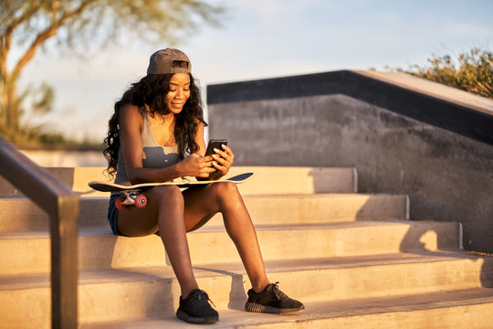 happy african american woman sitting on steps at skatepark with skateboard and smartphone