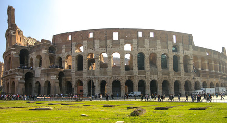 Naklejka premium Wide exterior of the Coliseum in Rome with clear morning skies and bright green grass, backlit by a lens flare