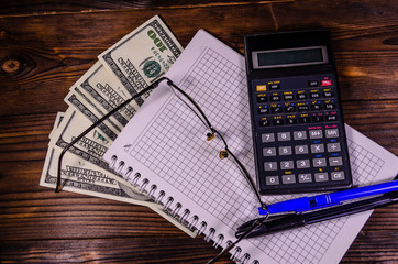 Notepad, pen, calculator and dollar bills on wooden table. Top view