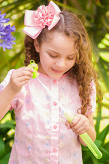 Happy little curly girl playing with soap bubbles on a summer nature, wearing a pink dress and flower in her head, in a blurred nature background