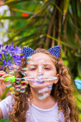 Happy little curly girl playing with soap bubbles on a summer nature, wearing a blue ears of tiger accessories over her head in a blurred nature background