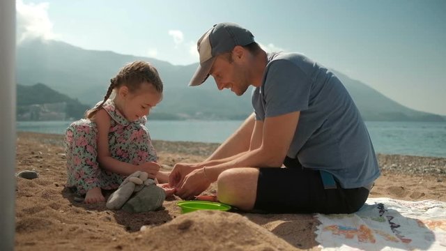 Young man with daughter sits on beach in summer day.