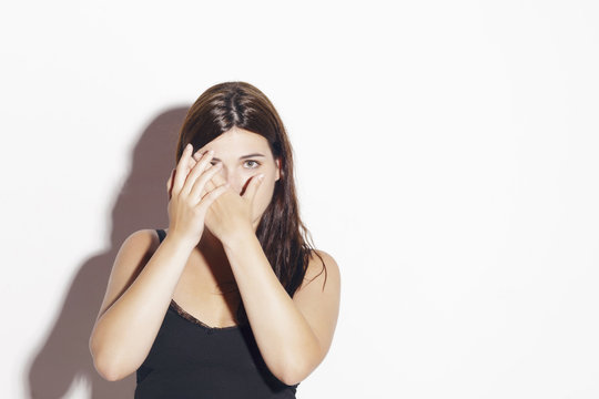 Pretty brunette girl hiding behind her hands, afraid of something. Fear. Indoors, over a white wall.