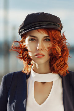 Outdoor close up portrait of young beautiful fashionable redhead woman posing in street. Model wearing stylish captain cap. Lady looking at camera. Female fashion concept. Sunny day light