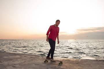 skater in red shirt and blue jeans riding on concrete pier on longboard during sunrise, sea or ocean background  