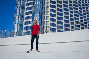 Young bearded man riding on skateboard, hipster with longboard in red 
shirt and blue jeans urban background  