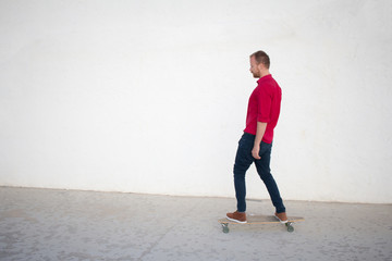 Young bearded man riding on skateboard, hipster with longboard in red 
shirt and blue jeans urban background  