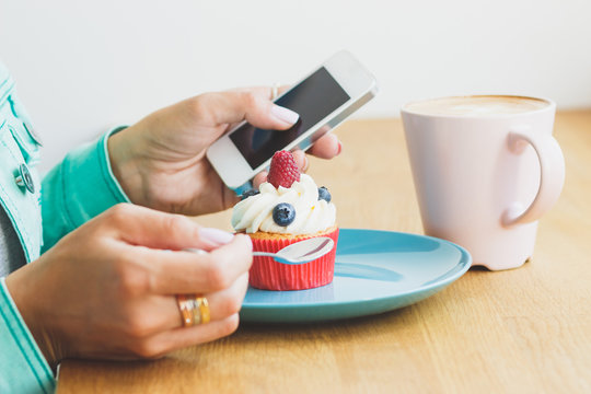 Woman takes a picture of cup with cappuccino and a cupcake on the plate