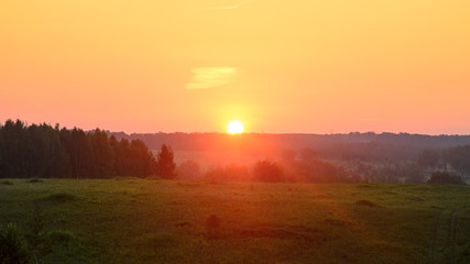 Bright yellow disk of the sun sits at the horizon of the forest and the towers of power lines against orange sunset rays in the fog on the slope of the landscape and the trees.