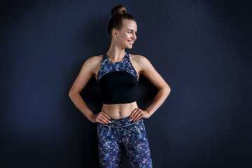 Beautiful fitness model is posing in front of the dark wall in a dark training suit