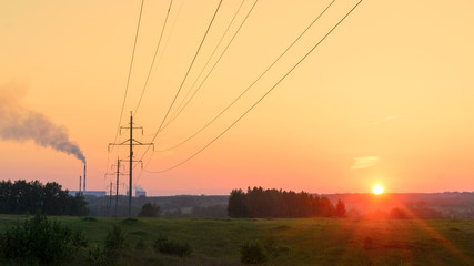 Pillars of power lines with wires standing in a row outside the city heat power plant with Smoking chimneys at sunset in a field on green grass next to the setting sun with rays and beautiful skies.