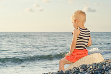 Fototapeta na wymiar Boy in striped vest sits on a rock by the sea. Back view