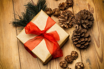 Fototapeta na wymiar Caja marrón con un lazo rojo junto a piñas y rama de pino sobre un fondo de madera rústico. Vista superior y de cerca. Concepto: Navidad, celebraciones
