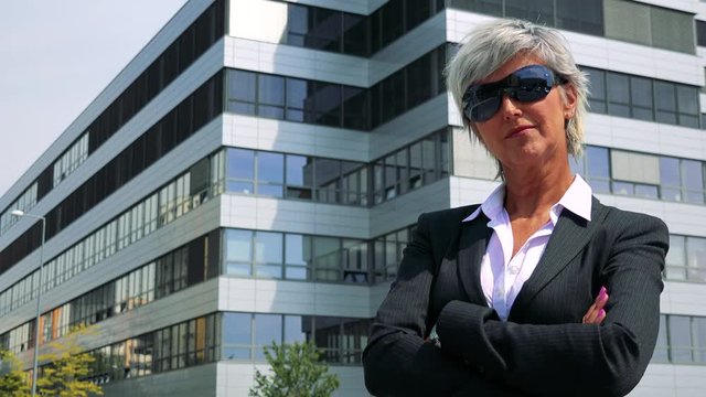 Business middle age woman puts on sunglasses and smiles to camera - company building in the background 