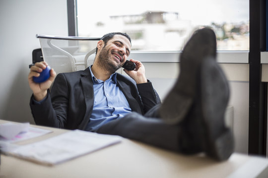 Manager seduto comodamente nel suo ufficio, con i piedi sopra la  scrivania parla divertito al telefono
