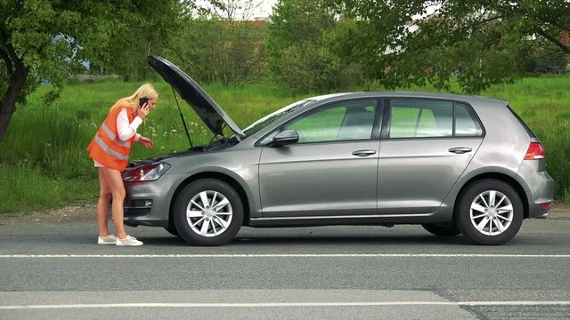Young attractive blond woman wears warning vest and phones with smartphone in front of broken car (open hood of the car) - cars passing in foreground - nature in the background 